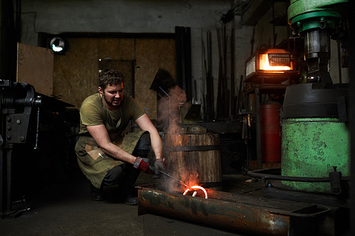 Young blacksmith in workwear putting hot molten metal workpiece into water to cool it down