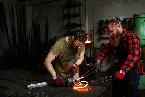 Two professional blacksmiths in workwear standing by anvil and processing hot iron workpiece in smithy