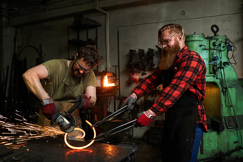 Serious skilled metalsmiths in aprons changing shape of twisted bar for fence: concentrated man cutting heated iron with drinder while long-bearded coworker holding it with tongs