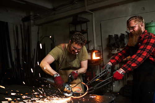 Concentrated manual workers in apron and safety goggles cutting heated twisted metal bar using grinder and standing at desk in workshop