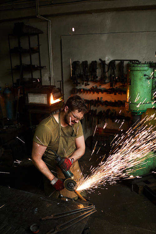 Serious busy strong male blacksmith in protective goggles grinding metal bar, sparkles blowing around