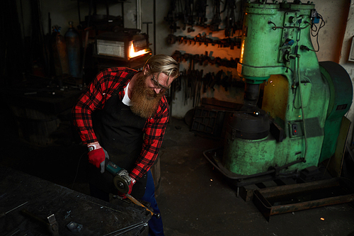 Serious concentrated brutal bearded worker in apron cutting metal with grinder while working in dark smithery