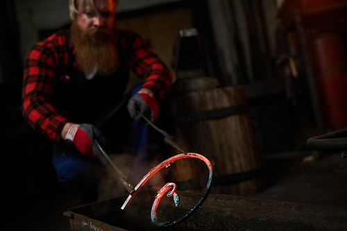 Serious brutal handsome blacksmith crouching while holding heated metal bar in tongs and putting it in water