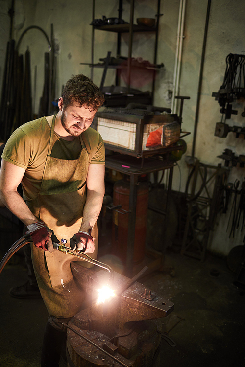 Young man in blacksmith apron and gloves using welding handtool while processing iron workpiece in smithy