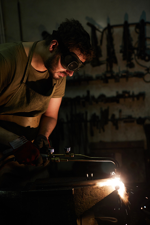 Serious concentrated bearded man in dark goggles using cutting torch while cutting steel detail on anvil in dark workshop, sparks blowing out