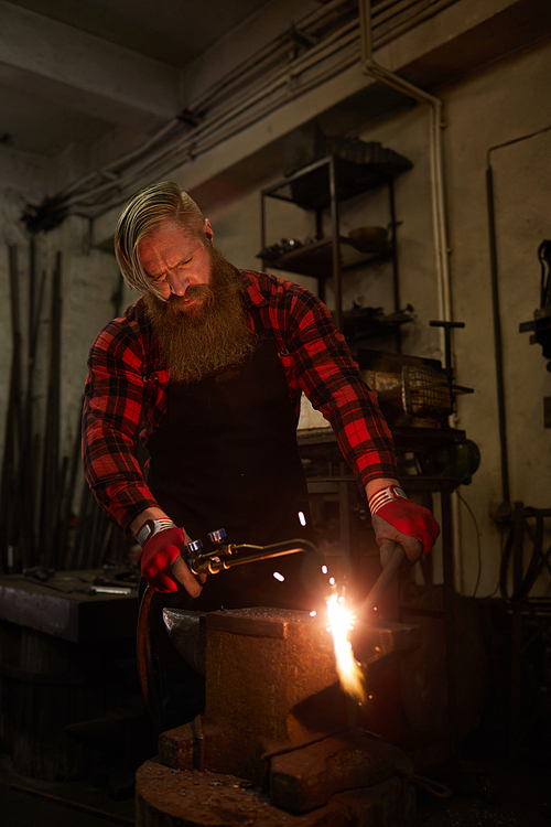 Serious brutal bearded blacksmith in apron and gloves concentrated on gas welding using torch while cutting steel bar in dark workshop