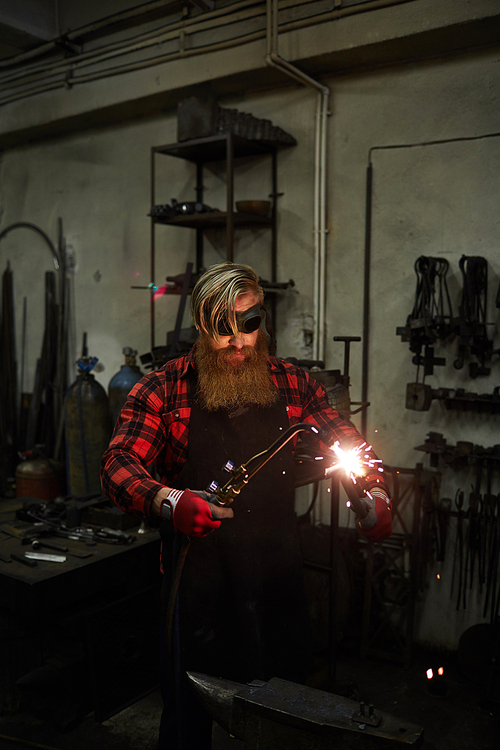 Serious handsome blacksmith with long beard wearing apron and checkered shirt holding steel part and cutting it with torch in workshop