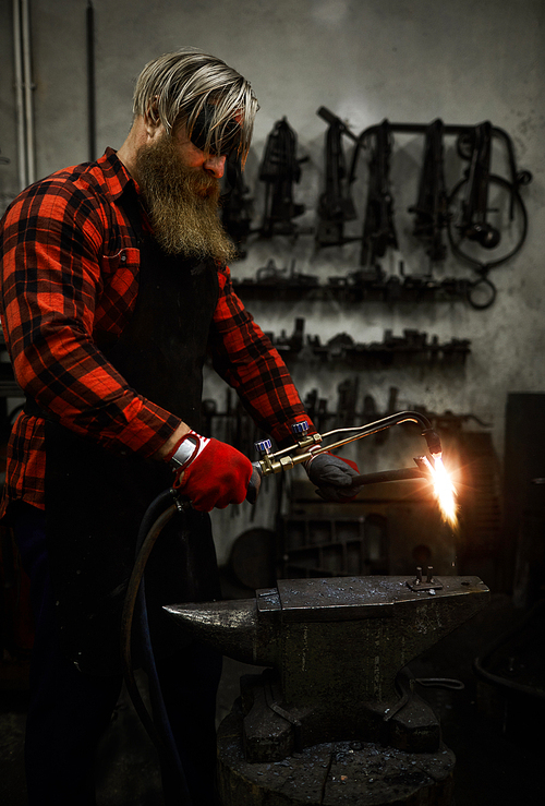 Bearded blacksmith in workwear working with welding handtool while standing by anvil