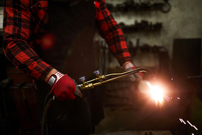 Close-up of unrecognizable smith in gloves using gas welding tool while cutting metal detail in workshop, sparkles blowing out during welding