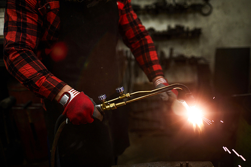 Close-up of unrecognizable smith in gloves using gas welding tool while cutting metal detail in workshop, sparkles blowing out during welding
