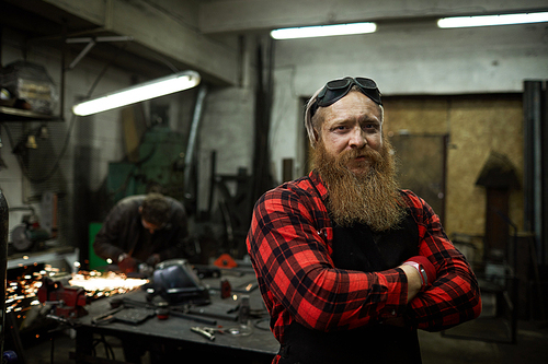 Content confident strong male blacksmith with long beard wearing safety goggles on head standing in welding shop and 
