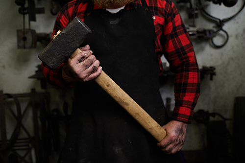 Close-up of unrecognizable smith in apron and checkered shirt holding big hammer in dirty hands, he ready for work