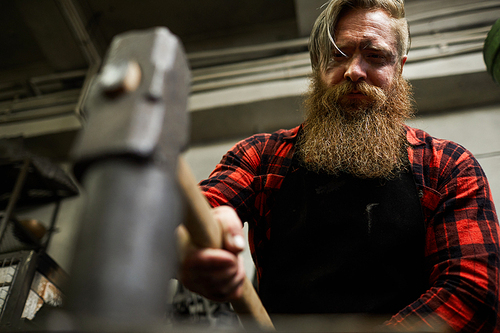 Serious frowning strong male blacksmith with long beard hammering metal detail while molding shape in workshop