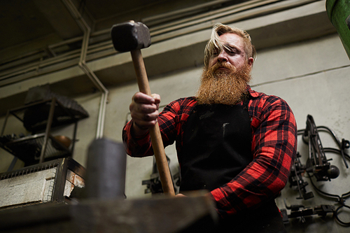 Blacksmith with beard holding big hammer over anvil while forging iron workpiece in smithy