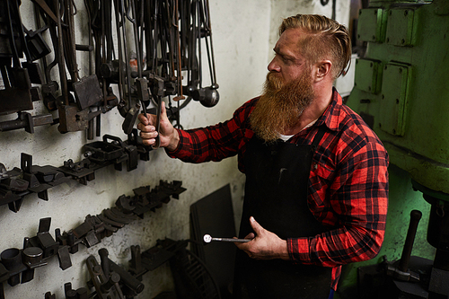 Serious busy blacksmith with long beard wearing apron and checkered shirt choosing tool for forging while preparing for work in workshop