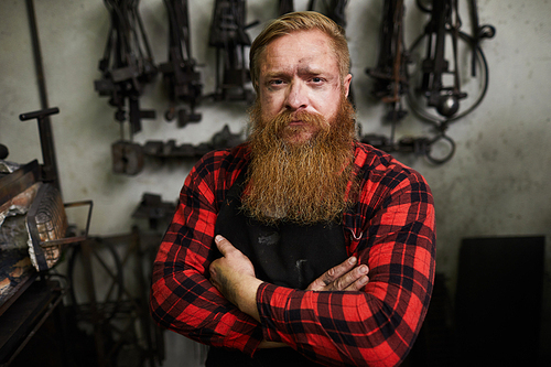 Portrait of serious confident handsome male smith with long beard looking scowling and crossing arms on chest in workshop