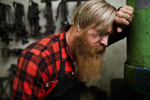 Exhausted male blacksmith with blond hair and long beard leaning on metal tube while standing in workshop and being without strength