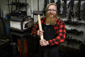 Positive confident strong male blacksmith in protective eyewear and apron holding big hammer for shaping metal and  in workshop