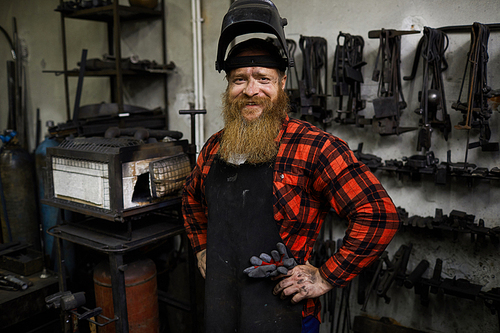 Cheerful satisfied handsome welder in mask on head and apron smiling at camera and holding hands on hips in workshop with various tools hanging on wall