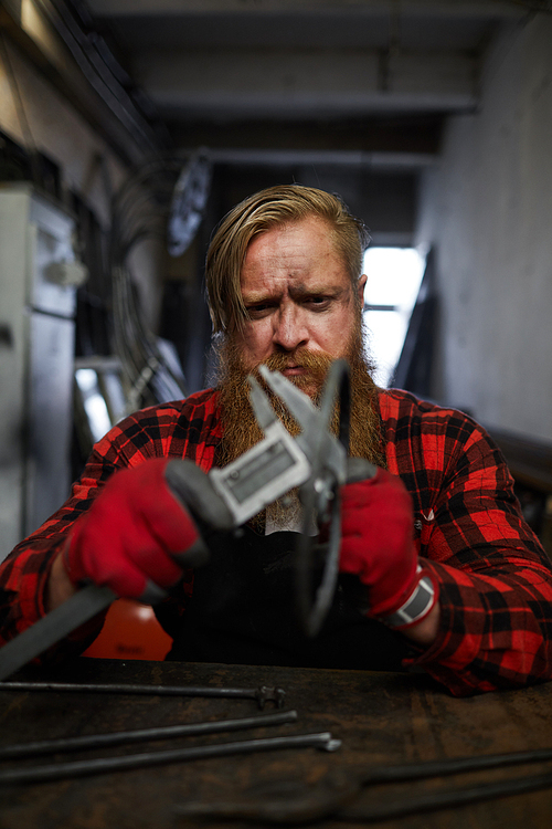 Frowning concentrated brutal worker with beard measuring steel detail using vernier caliper while sitting at table in workshop