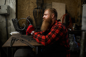 Sad concentrated male blacksmith with long beard checking twisted metal bar made by hands in workshop