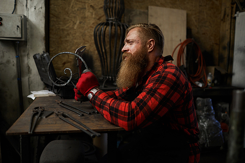 Sad concentrated male blacksmith with long beard checking twisted metal bar made by hands in workshop