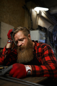 Sad knackered long-bearded manual worker in checkered shirt and gloves sitting at table and leaning on hand in workshop