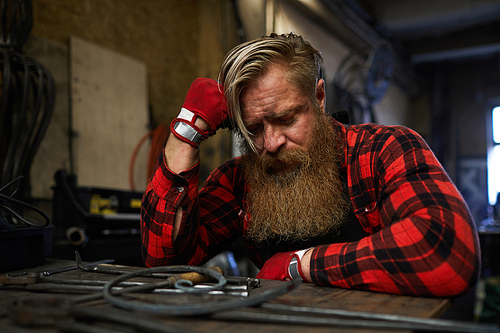 Tired brutal craftsman with long beard leaning on hand and sleeping at table after overworking