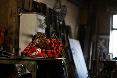 Tired male blacksmith in checkered shirt and gloves sleeping at table and keeping head on arms in workshop, overworking concept