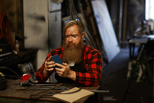 Frowning puzzled bearded blacksmith in checkered shirt using smartphone while checking message and sitting at table with hand tools in workshop