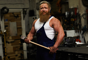 Content athletic handsome blacksmith in overall wearing protective eyewear on head holding heavy hammer and  in workshop