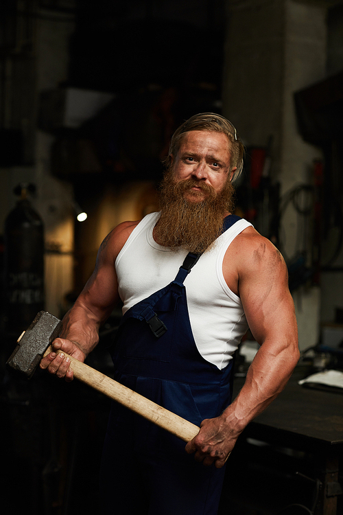 Serious brutal strong man in workwear holding hammer and  in dark smithery, muscular worker in workshop