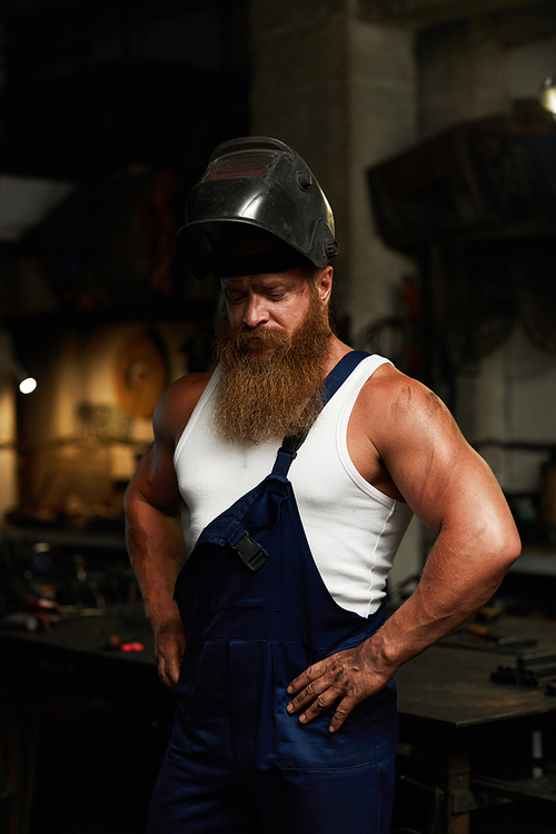 Serious dirty handsome muscular welder in mask looking down and holding hands on hips in dark workshop