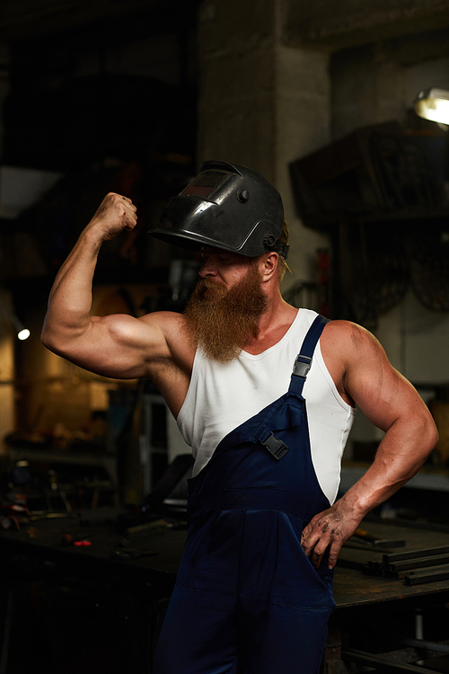 Serious athletic welder in open mask flexing bicep and looking at it while showing his power in workshop