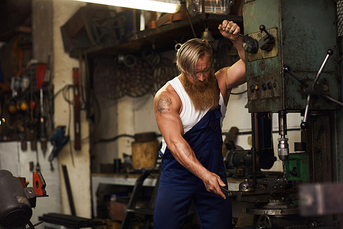Serious powerful handsome blacksmith with tattoo on shoulder adjusting drilling machine while working in own smithery