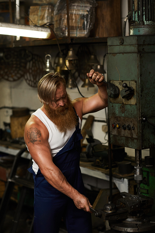 Serious concentrated muscular craftsman with tattoo and long beard working with drilling machine and using handles to fix detail