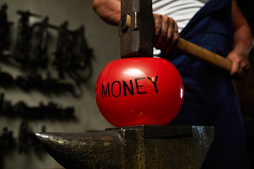 Red ball symbolizing money concept is situated on anvil with big heavy hammer above