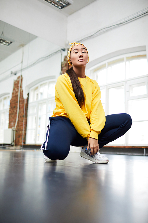 Young active female in sweatshirt, sweatpants and sneakers squatting in modern dance studio