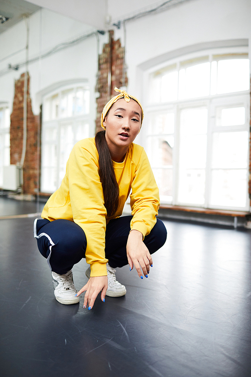 Female dancer in activewear squatting on the floor of modern dance studio and looking at you
