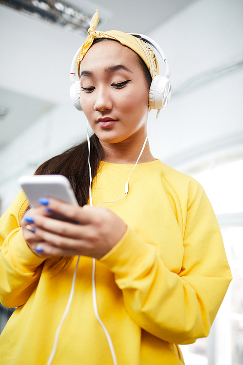 Active girl in yellow sweatshirt and headband listening to music in headphones while choosing soundtrack from playlist