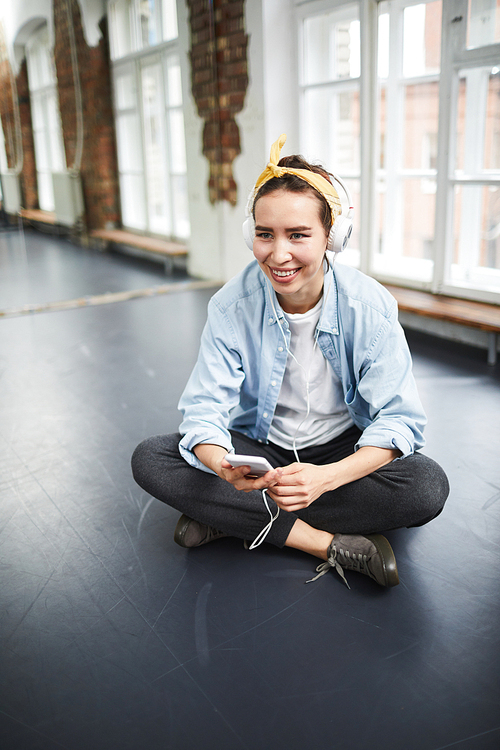Happy young cross-legged woman in activewear sitting in studio of dance and listening to music at break