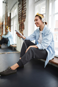 Young restful woman in headphones listening to music at break after breakdance training