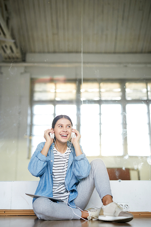 Happy girl putting earphones into ears while sitting on the floor of modern dance studio at break