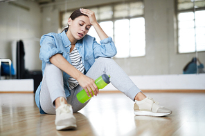 Exhausted active woman with bottle of water sitting on the floor of modern dance studio and having break