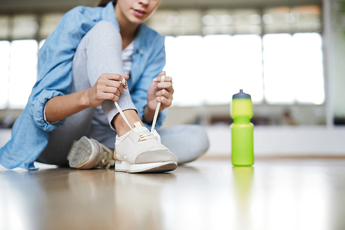 Young active female tying shoelaces of sneakers while sitting on the floor with plastic bottle near by