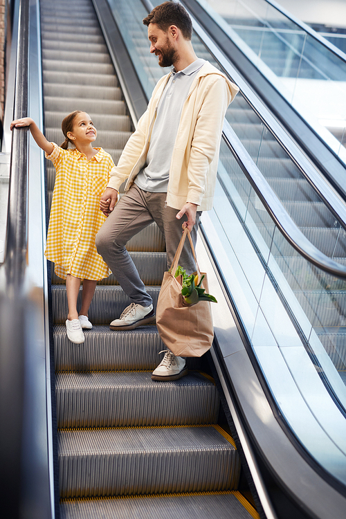 Positive bearded father and pretty daughter holding hands and chatting while moving down escalator in mall, handsome father holding bag full of fresh products
