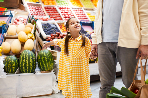 Smiling pretty girl in checkered dress pointing at watermelon and asking father to buy it in fresh food market, they holding hands while walking over store