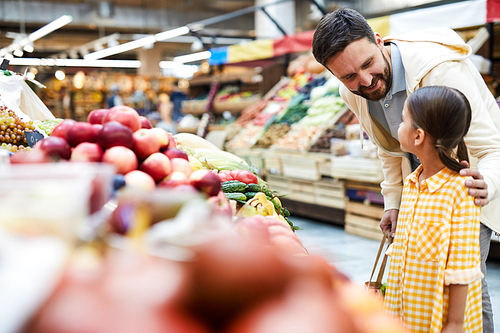 Cheerful careful father with beard touching shoulder of daughter and asking her about food preferences while they choosing organic food at farmers market