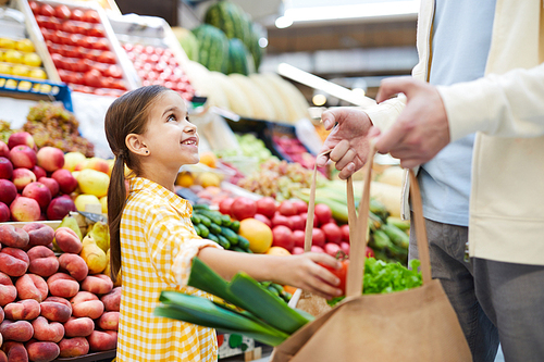 Cheerful cute girl in yellow dress putting vegetable into bag and looking at father while helping him with shopping at farmers market