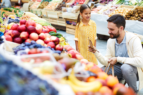 Content handsome young bearded father crouching near food stall and checking shopping list at farmers market while buying organic food together with daughter